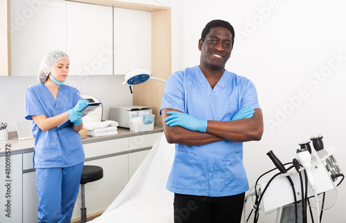 Portrait of confident african american male cosmetologist standing in modern aesthetic medicine office on background with female assistant filling out medical form..