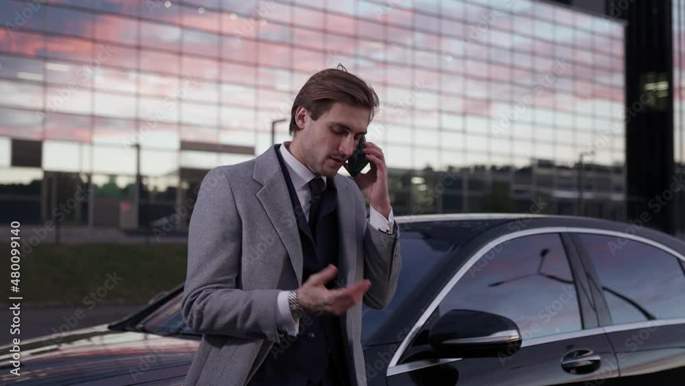 Handheld, portrait of a elegant businessman standing near a business car and talking on a mobile phone, top manager in a suit, evening time, sunset. Handheld, portrait of a elegant businessman standing near a business car and talking on a mobile phone, top manager in a suit, evening time, sunset.