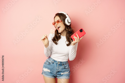 Summer girl laughing, eathing lolipop and listening music in headphones, dancing with smartphone against pink background photo