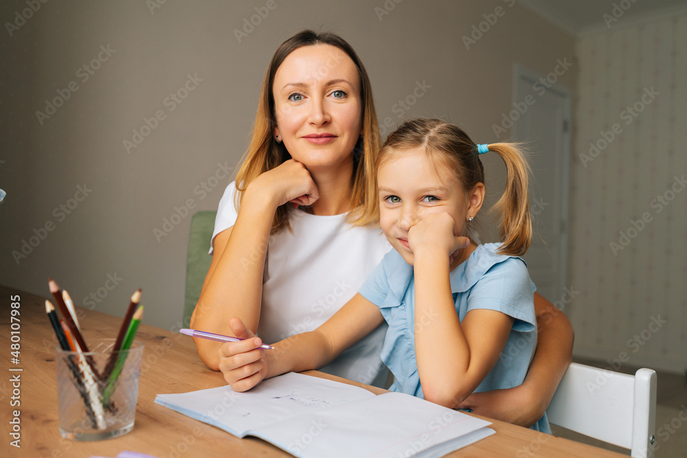 Close-up of happy elementary daughter doing homework with young mother sitting at home table by window, looking at camera. Smiling female tutor teaching elementary school girl helping with lesson. Close-up of happy elementary daughter doing homework with young mother sitting at home table by window, looking at camera. Smiling female tutor teaching elementary school girl helping with lesson.