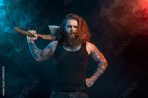 Fototapeta Naklejka Na Ścianę i Meble -  Handsome man with cigar holds old axe. Bearded lumberjack. Serious male with tattoos with long hair.
