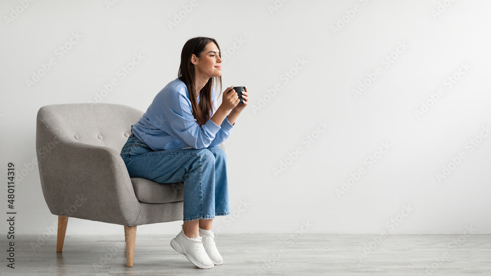 Time for coffee. Charming millennial woman sitting in armchair, enjoying cup of hot drink against white wall Time for coffee. Charming millennial woman sitting in armchair, enjoying cup of hot drink against white wall