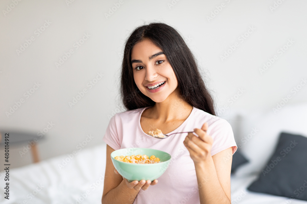 Charming Indian lady eating tasty cereal with milk, enjoying nutritious cornflakes in bedroom