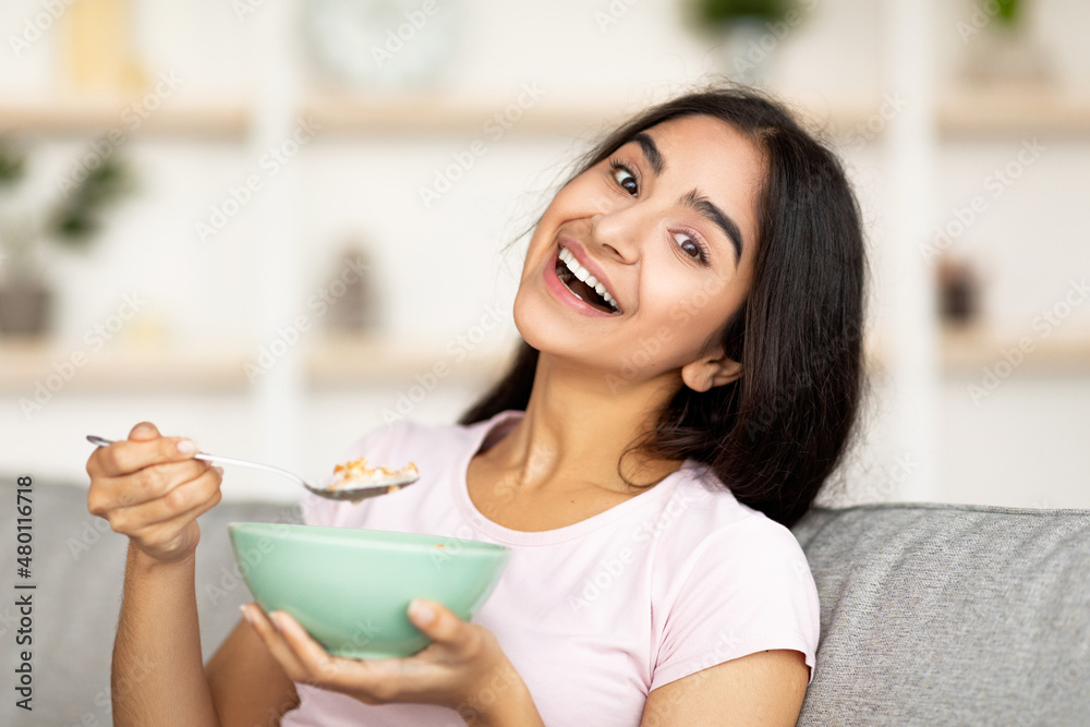 Joyful Indian lady eating granola or cereal with milk on comfy couch at home, smiling at camera Joyful Indian lady eating granola or cereal with milk on comfy couch at home, smiling at camera