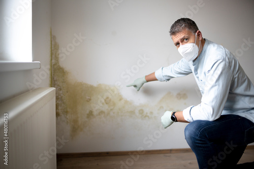 Man with mouth nose mask and blue shirt and gloves n front of white wall with mold photo