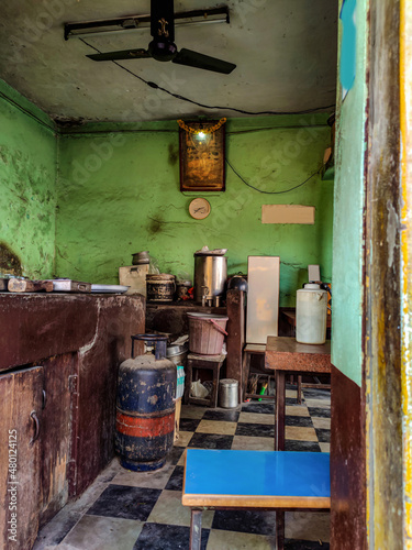Inside view of Old Indian dirty abandoned hotel kitchen, gods photo and clock hanging on pistachio color painted wall. Gas cylinder, old utensils, wooden table , chair and other objects in the kitchen © Vinodkumar