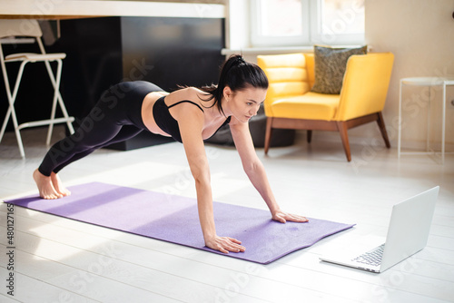 Attractive woman in black sport clothes training on yoga mat and watching tutorial on modern laptop. Concept of domestic workout.