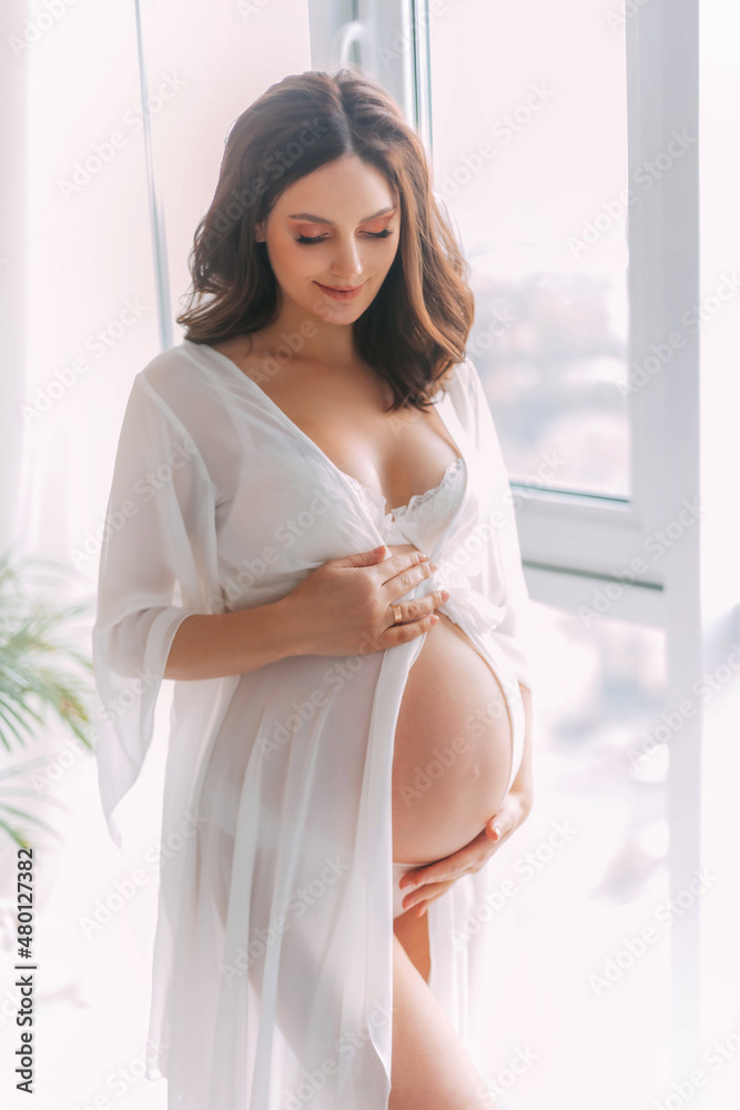 Young beautiful pregnant woman stand near window hugs bare belly with  hands. White long silk negligee pregnancy dress peignoir, dressing gown.  Light room classic background. Gently smiling face Stock Photo