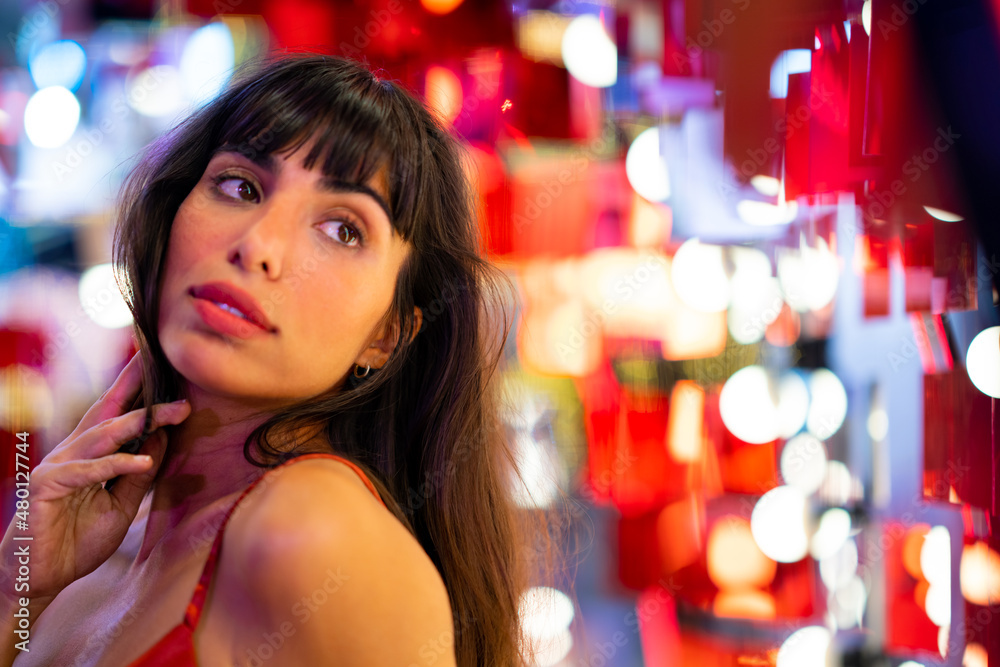 Portrait of Beautiful Hispanic woman in red dress enjoy with illuminated festive lights in the city. Confidence female having fun with glowing decorations night lights of holiday festival celebration.