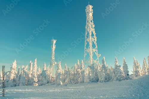Cell towers and fir trees covered with snow on top of a mountain on a winter day. Blue sky in the background.