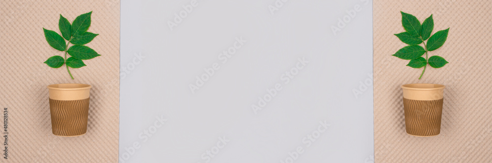 Banner with copy space. Mockup image of eco-friendly coffee to go cup - kraft paper cups with green leaves above on light gray background. Sustainable packaging and zero waste concept. Selective focus Banner with copy space. Mockup image of eco-friendly coffee to go cup - kraft paper cups with green leaves above on light gray background. Sustainable packaging and zero waste concept. Selective focus