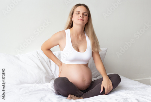 Beautiful blonde pregnant woman is sitting on the bed. Experiencing back pain. Problems bearing a child. Close-up. The background.