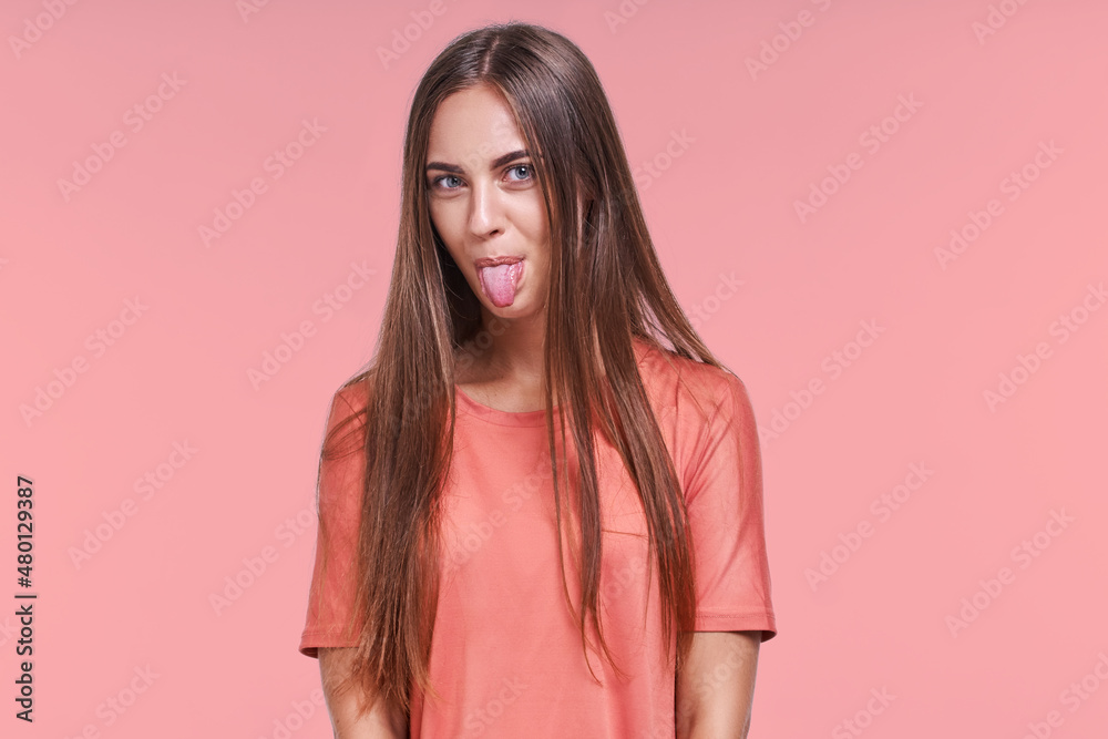 Funny pretty young Caucasian woman with long hair makes comic face showing tongue having fun alone wears casual t-shirt standing on studio pink background. Lifestyle, face expressions Funny pretty young Caucasian woman with long hair makes comic face showing tongue having fun alone wears casual t-shirt standing on studio pink background. Lifestyle, face expressions