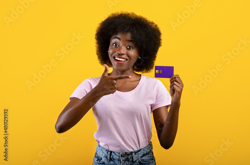 Positive african american bank customer showing credit card, recommending bank services, yellow background