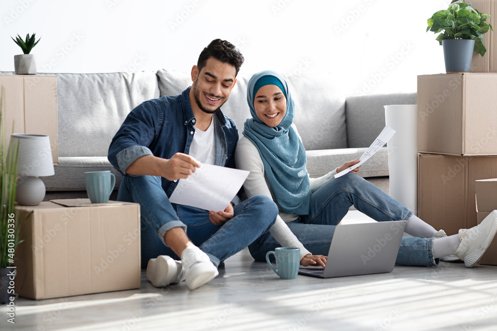 Smiling muslim family sitting on floor with laptop among boxes Smiling muslim family sitting on floor with laptop among boxes