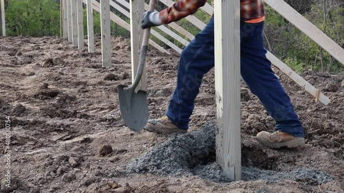 Construction worker using shovel to put cement into post hole for principal pole of new barn construction; uses his foot to level and tamp cement; concepts of new construction and blue collar labor photo