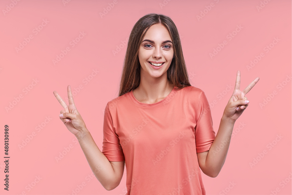 Cheerful young pretty caucasian woman with smile shows victory gesture with two hands and looks directly at you to greet, carefree posing on pink background in studio. positive life style Cheerful young pretty caucasian woman with smile shows victory gesture with two hands and looks directly at you to greet, carefree posing on pink background in studio. positive life style
