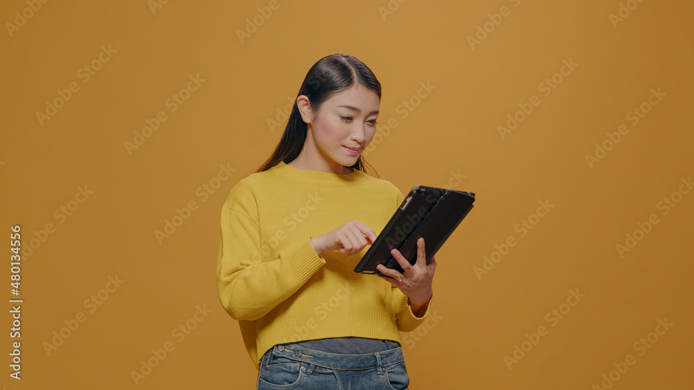 Focused model browsing internet on digital tablet in studio, using device with touch screen to do successful work. Asian woman holding modern gadget with app connection over background. Focused model browsing internet on digital tablet in studio, using device with touch screen to do successful work. Asian woman holding modern gadget with app connection over background.