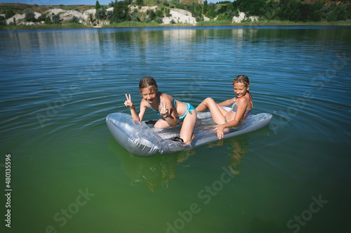 Two little cute girls in swimsuits swim on an inflatable mattress on a blue career lake. © bearok