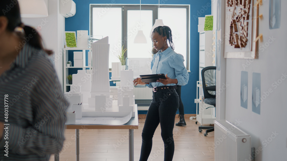 African american woman using digital tablet to design blueprints plan with building model on table. Industrial engineer working with maquette, planning construction layout and structure.