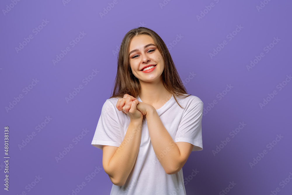 Portrait of smiling positive woman keeps hands together expresses her feelings to girlfriends, shows love and great sympathy, wearing blank white t-shirt, standing over purple background