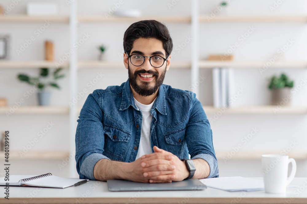 Happy toung arab self-entrepreneur man sitting at desk in home office interior and smiling to camera Happy toung arab self-entrepreneur man sitting at desk in home office interior and smiling to camera