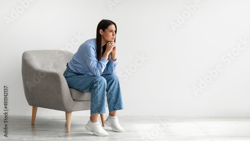 Tableau sur toile Portrait of annoyed young lady sitting in armchair against white studio wall, ba