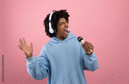 Creative Afro teen in headphones listening to music, singing karaoke song into microphone on pink background photo