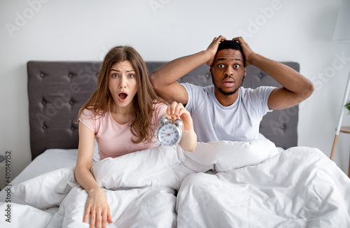 We are late. Shocked interracial couple with alarm clock grabbing their heads in terror, sitting on bed at home
