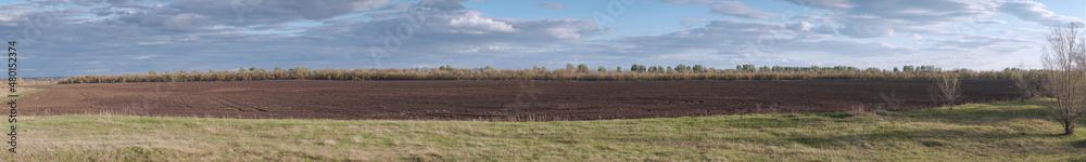 Panorama of a plowed agricultural field under a cloudy sky in front of a forest