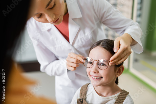 ophthalmologist trying eyeglasses on smiling girl near blurred mom in optics store.