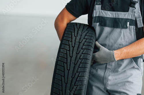 Man in uniform is working in the autosalon at daytime. Holding tire in hands