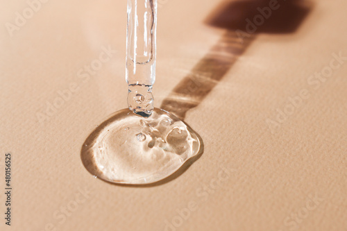 A drop of skin moisturizing serum flowing from a pipette on a beige neutral background close-up.