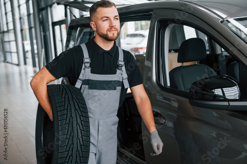 With tire in hands. Man in uniform is working in the autosalon at daytime photo