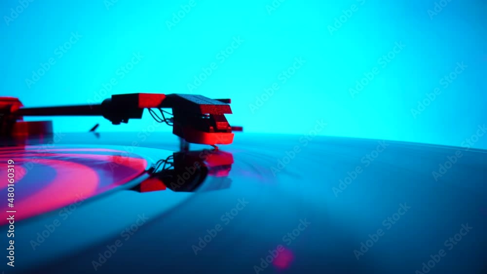 Black vinyl record spinning and play music on old turntable in rays of bright red and blue neon lights. Turntable needle on vinyl record, reads recording from grooves on disc. Close up. Slow motion.