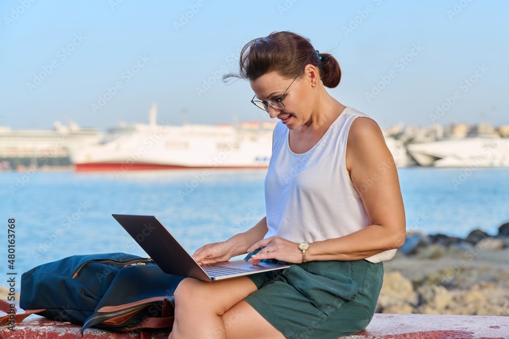 Middle-aged woman tourist with a backpack in seaport using a laptop