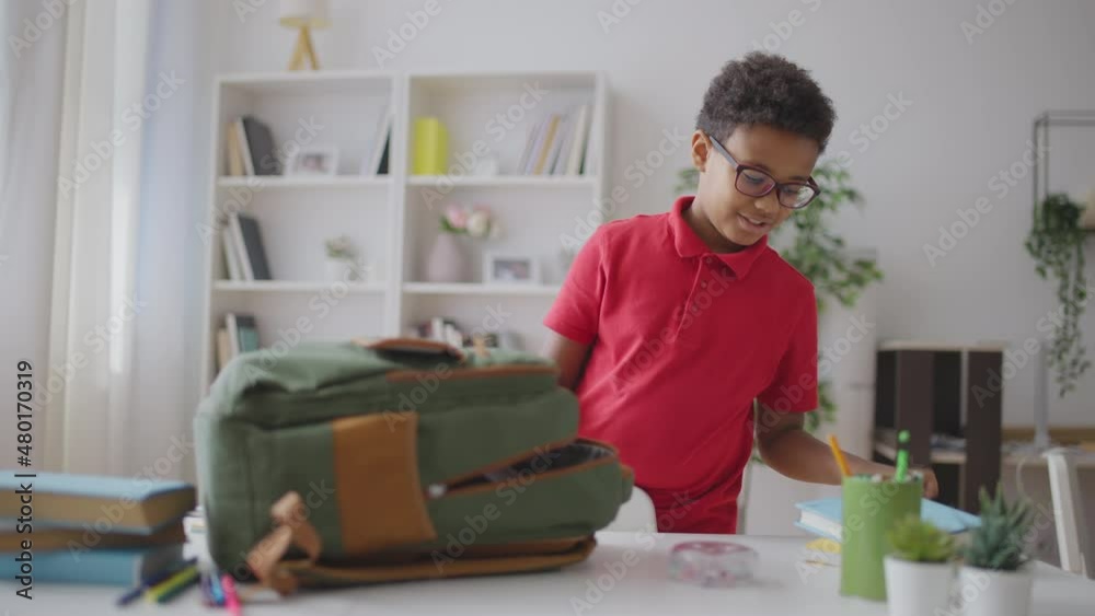 Pupil in eyeglasses packing backpack for school, putting books inside, routine Pupil in eyeglasses packing backpack for school, putting books inside, routine