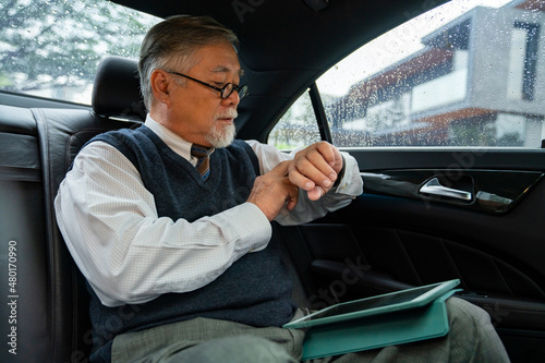 Confidence senior businessman CEO in suit sitting on car backseat working on digital tablet with checking time on hand watch while going to office. Elderly businessman and transportation concept. © CandyRetriever 