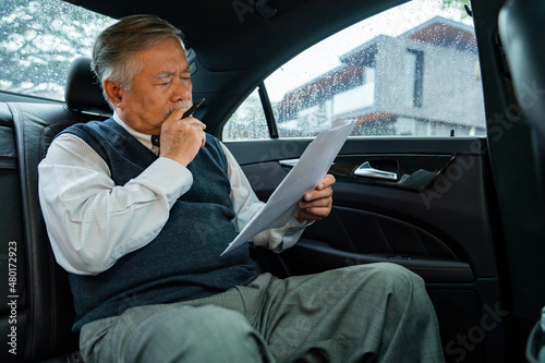 Confidence senior businessman in suit sitting on car backseat and reading business plan document while going to office. Elderly CEO working in automobile and looking at traffic out of the window © CandyRetriever 