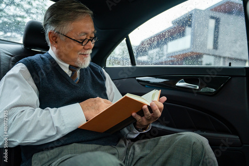 Confidence senior businessman CEO in suit wearing glasses sitting on car back seat and reading a book while going to work at office. Elderly businessman and transportation concept. © CandyRetriever 