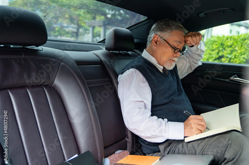 Confidence senior businessman CEO in suit wearing glasses sitting on car back seat and writing business plan in the book while going to work at office. Elderly businessman and transportation concept.