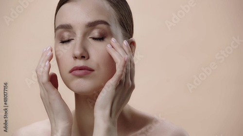 young woman with bare shoulders and closed eyes applying face cream isolated on beige.