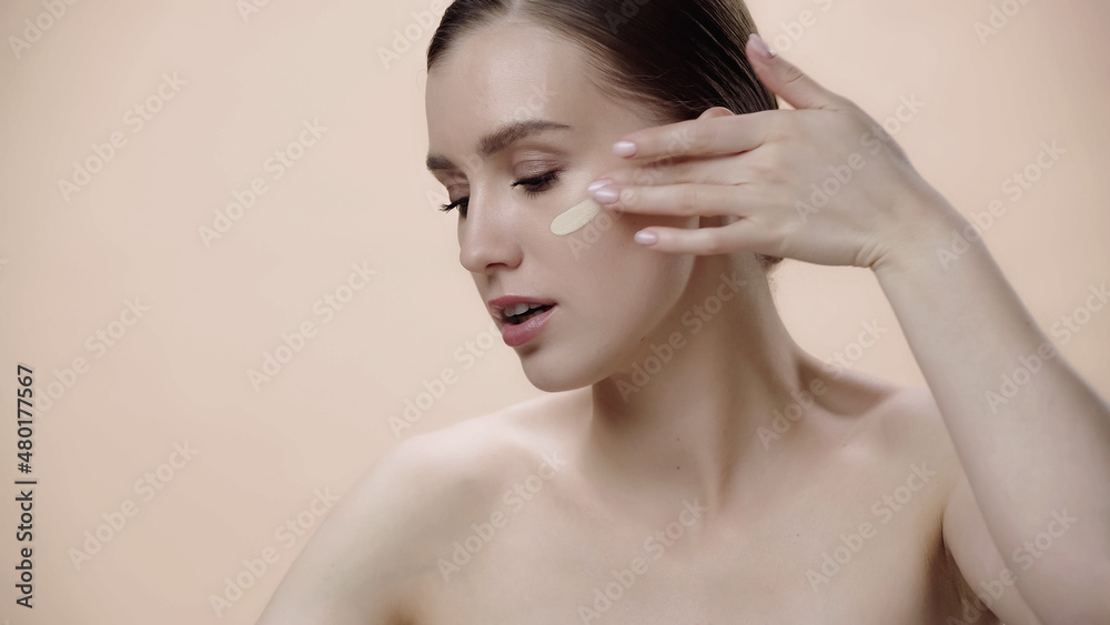 pretty young woman with bare shoulders applying face foundation isolated on beige.