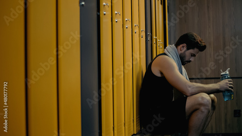 Konstfotografi side view of sportsman sitting on bench and holding sports bottle in locker room