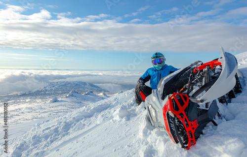 close-up of a powerful snowmobile piled on its side and a rider sitting on top of a mountain. a sports snowmobiler in bright clothes and a helmet without brands, a colorful snowmobile of a new model
