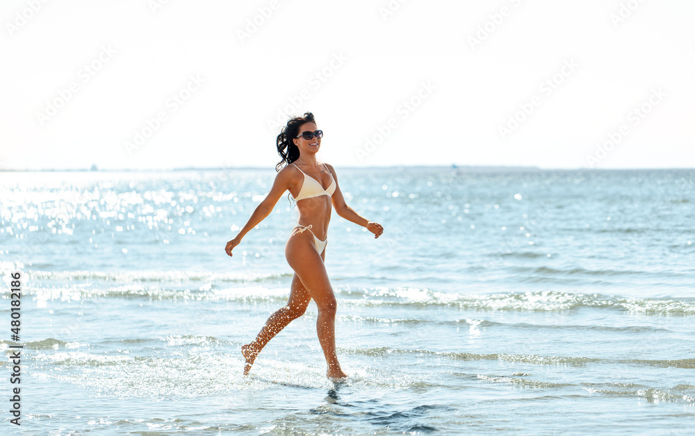 people, summer and swimwear concept - young woman in bikini swimsuit running in shallow water on beach