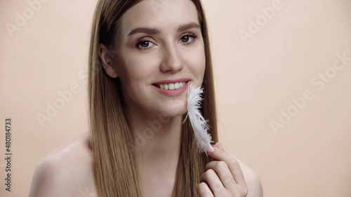 happy young woman with bare shoulders touching face with white feather isolated on beige.