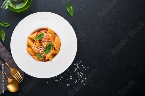 Bolognese Penne Pasta. Tasty appetizing classic italian penne pasta with parmesan, basil and Bolognese sauce in white plate on plate on black dark background.Traditional Italian cuisine. Top view.