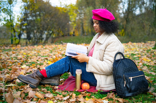 Serious dark-skinned intelligent african american woman, beautiful stylish ethnic girl reader reading a book in a golden autumn park in autumn, sitting on the grass