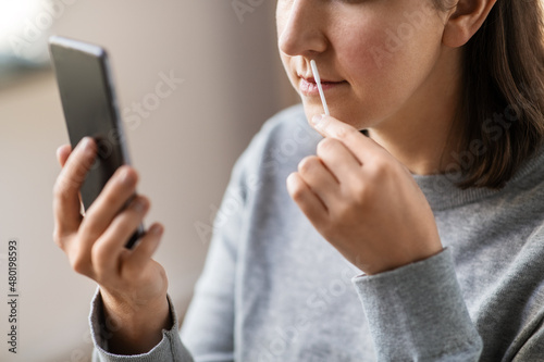 medicine, quarantine and pandemic concept - close up of woman with swab and smartphone taking sample from her nose and making nasal coronavirus self test at home photo